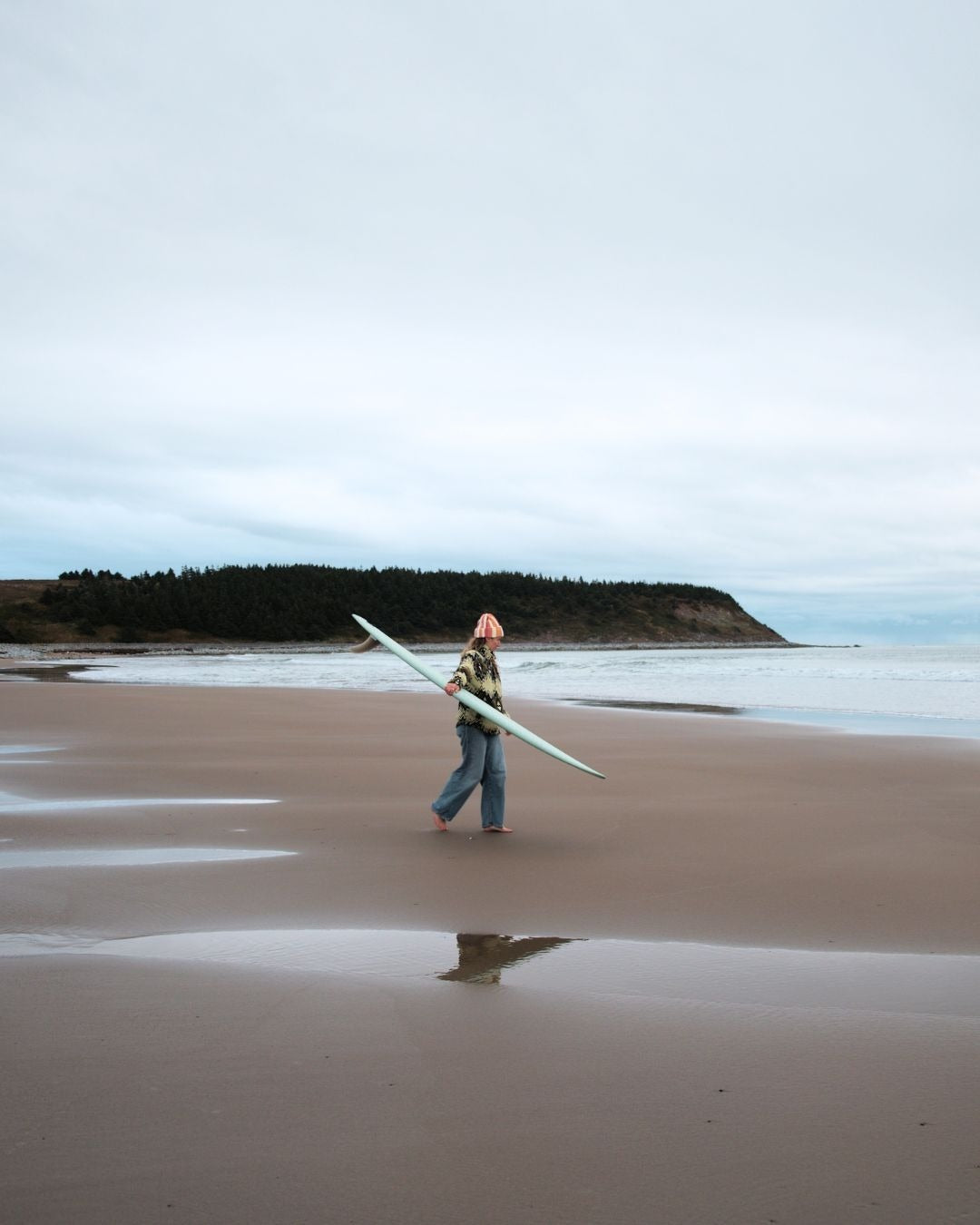 Person walking on a beach holding a surfboard with a scenic background