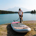 Man inflating a paddleboard on a lakeside with mountains in the background