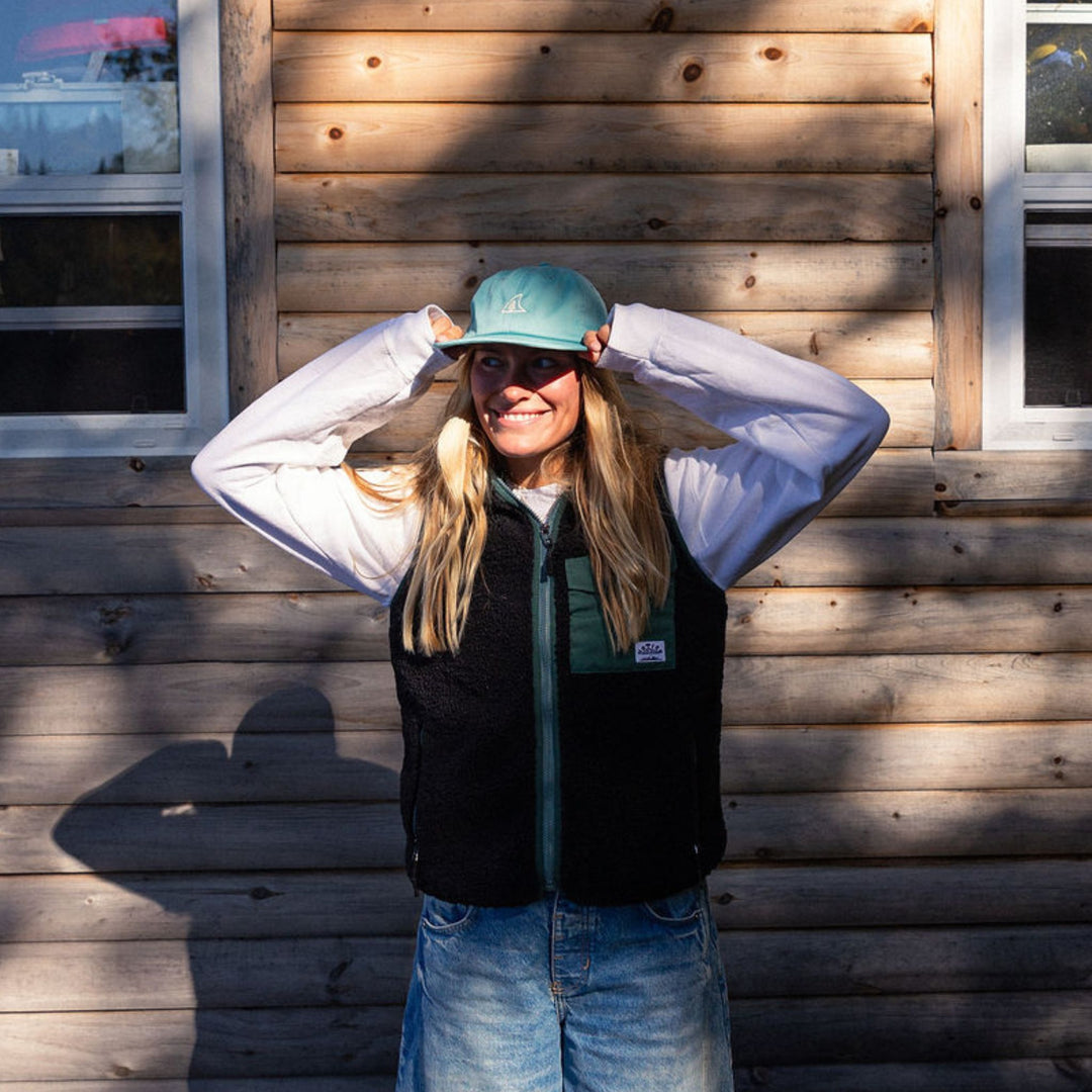 Person wearing a blue cap and black vest standing against a wooden wall.