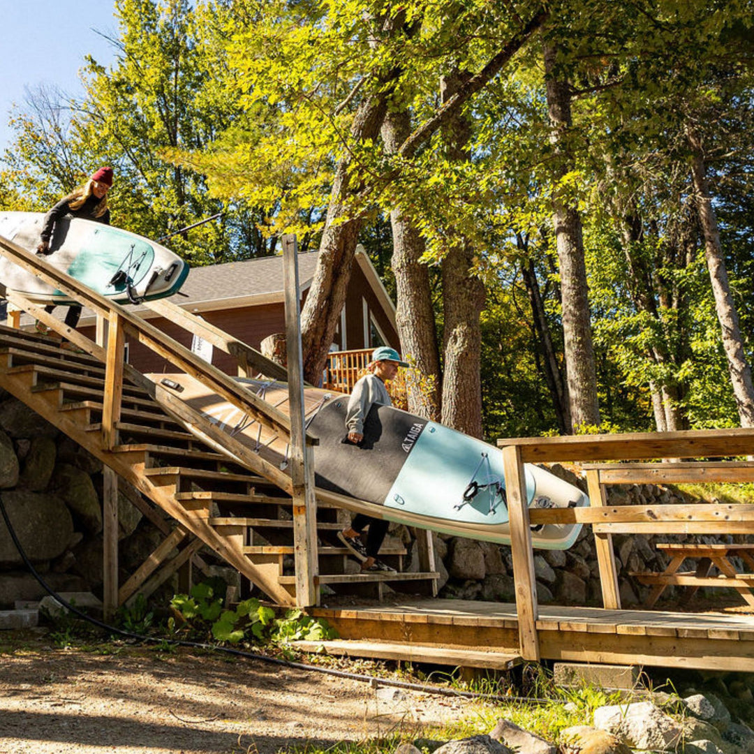Two people carrying paddleboards down a wooden staircase in a forested area.