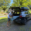 Man carrying a surfboard in its surf sock bag in an open car trunk with a roof rack.
