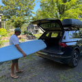 Man carrying a surfboard in its surf sock bag in an open car trunk with a roof rack.