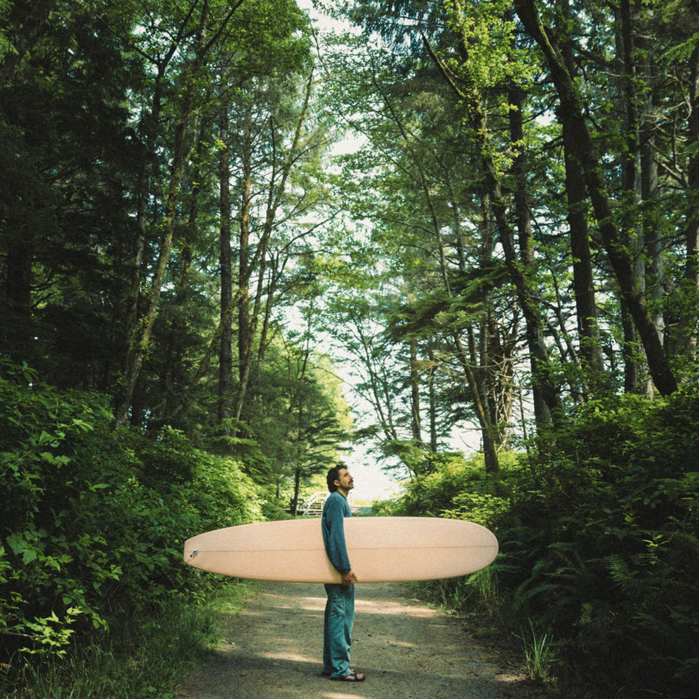 Guy Holding The Longboard Surf Board in The Rosé Color In The Forest