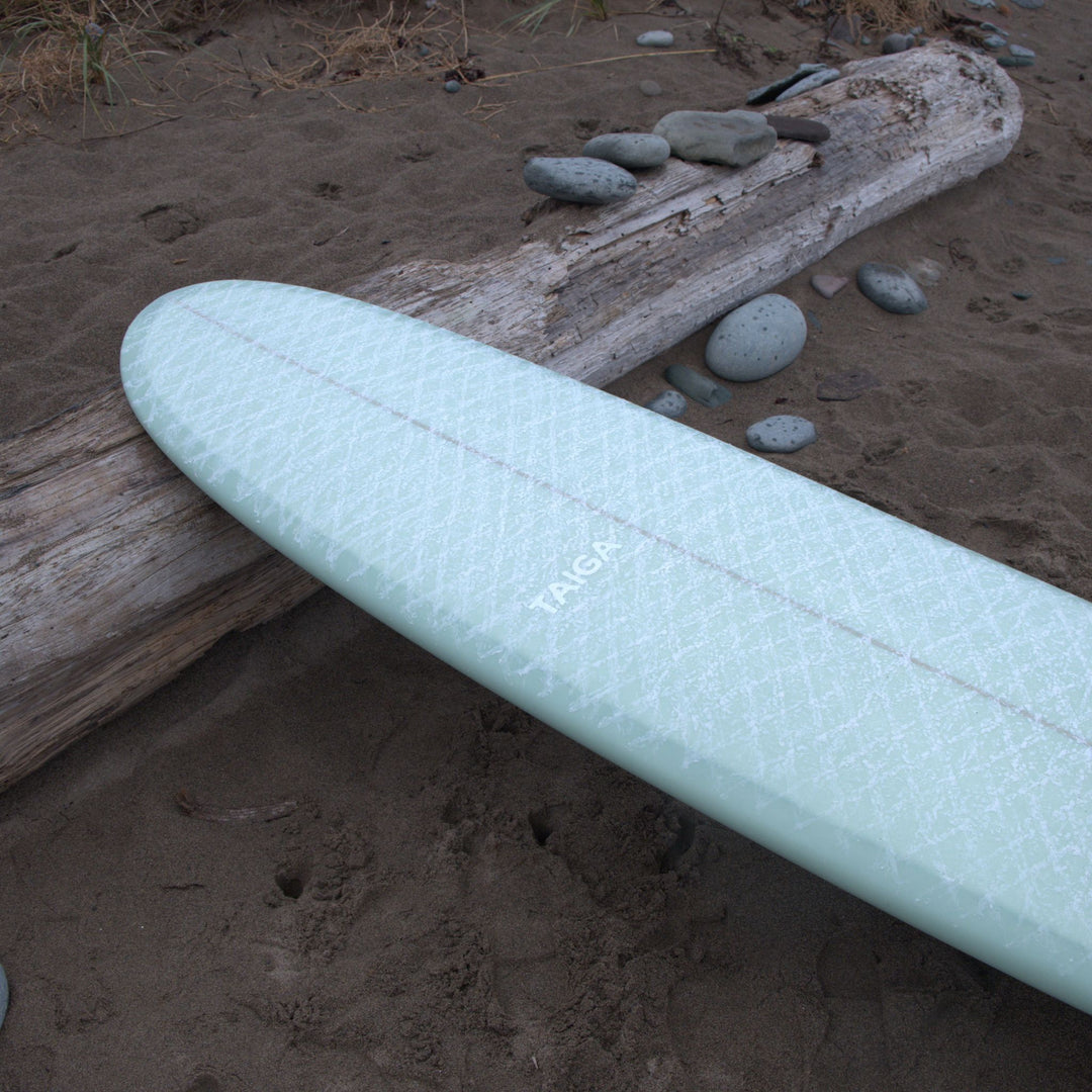 Sage Taiga surfboard on a sandy beach with driftwood and stones.