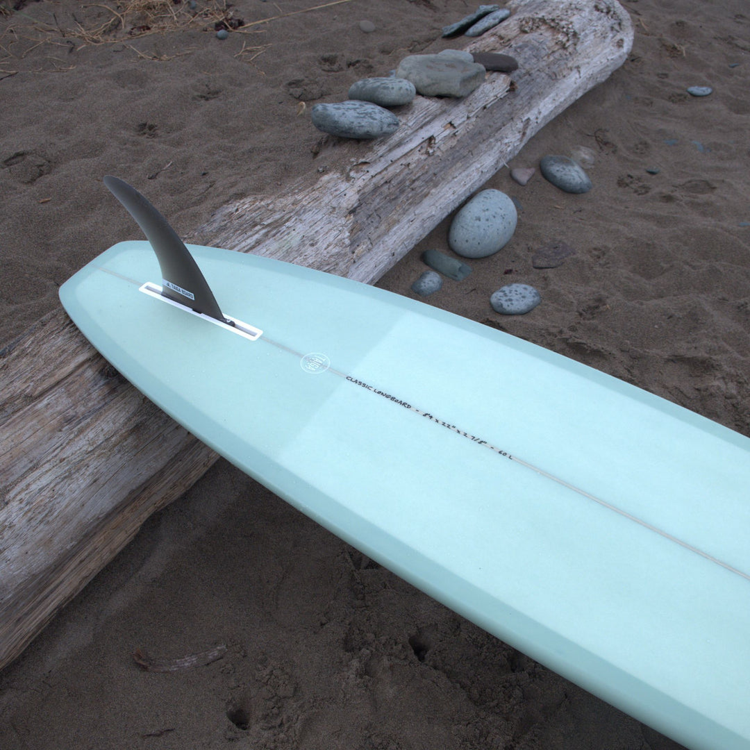 Sage surfboard on a sandy beach with driftwood and stones.