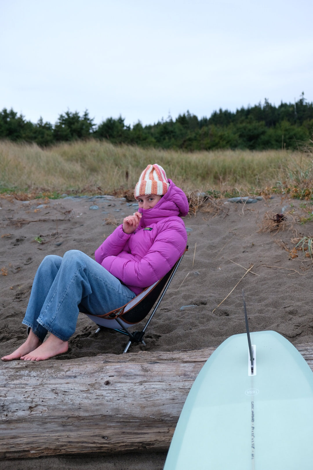Person sitting on a beach chair with a pink jacket and striped hat, next to a TAIGA Dusty Sage Longboard surfboard.
