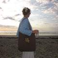 Person standing on a beach with a brown tote bag, holding a drink, against a sunset sky.