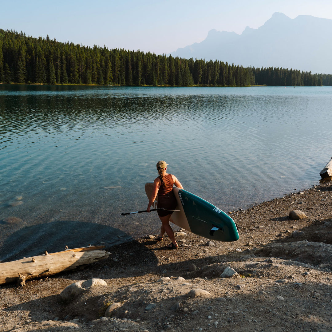 Person carrying a paddleboard on a lakeside with mountains in the background