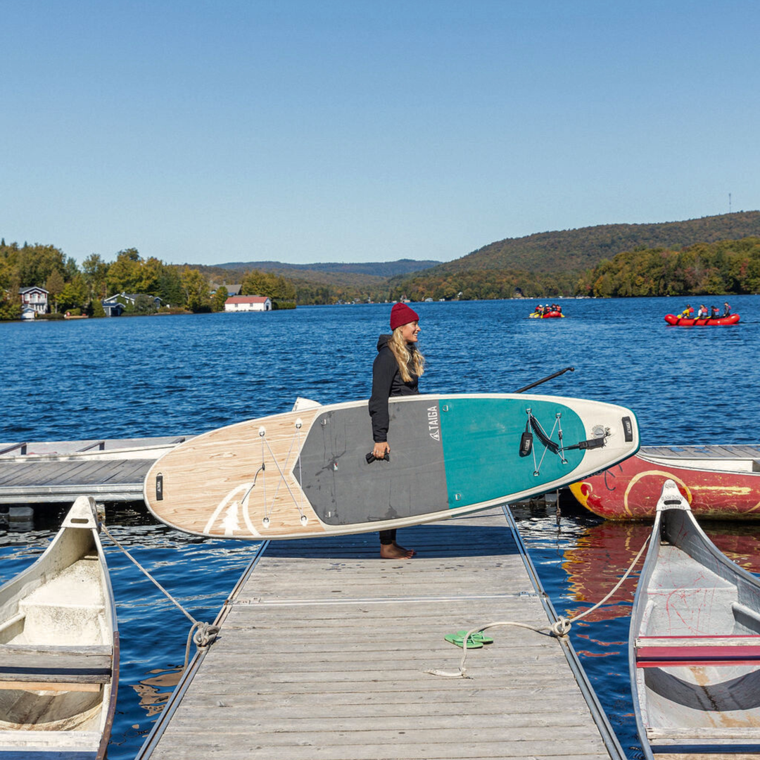 Person carrying a paddleboard on a dock with a lake and mountains in the background
