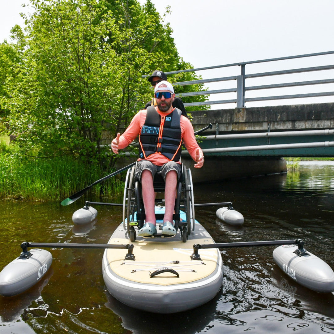Two people on a paddleboard with one in a wheelchair, on a calm body of water.