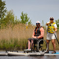 Two people on a paddleboard, one in a wheelchair, with nature in the background