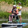 Man in a wheelchair and woman on a paddleboard with reeds and trees in the background