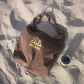Brown tote bag with 'TAIGA BOARD' logo on sandy ground